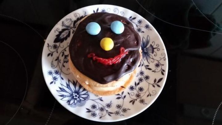 Ein mit Schokolade &uuml;berzogener Schaumkuss zeigt ein Gesicht mit bunten Smarties als Augen und Nase, auf wei&szlig;em, blumigem Teller.