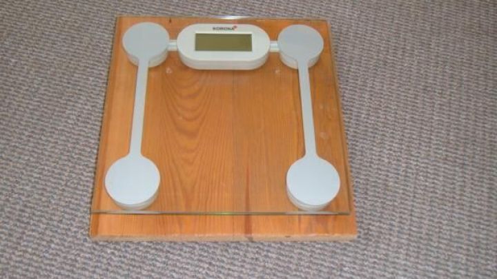 Digitale Personenwaage zeigt Gewicht, umgeben von Holzboden und Teppich. Text auf Waage: "SCRENNI".