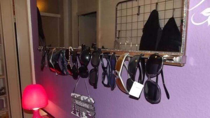 Eine Reihe von Sonnenbrillen h&auml;ngt ordentlich an einem Gitter &uuml;ber einer lila Wand, neben einer Lampe und einem Spiegel.