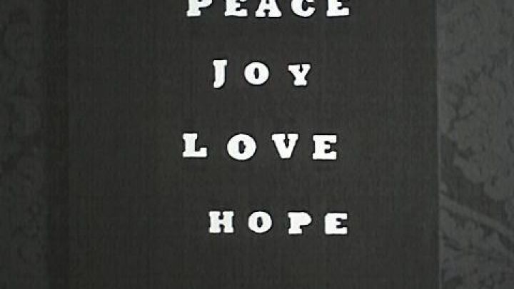 Leinwandbild mit den strahlenden Worten "PEACE JOY LOVE HOPE" leuchtet vor schwarzem, gemustertem Hintergrund.