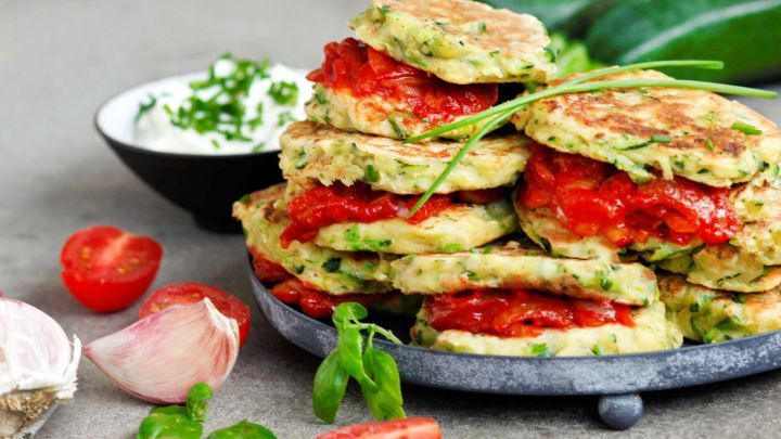 Zucchini-Pfannkuchen t&uuml;rmen sich mit Paprika-F&uuml;llung auf einem Teller, umgeben von frischen Kr&auml;utern und Tomaten.