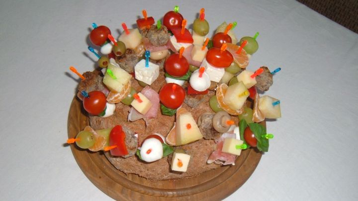 Ein Brotigel steckt auf einem Holzteller voller Party-Spie&szlig;chen mit Tomaten, K&auml;se, und Trauben in einer Feierumgebung.