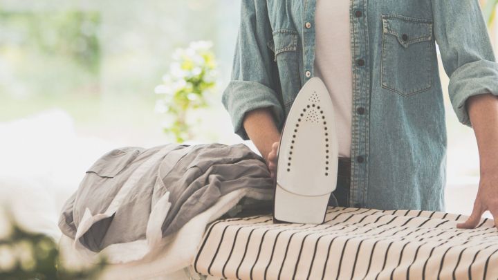 Ein B&uuml;geleisen steht auf einem Tisch, w&auml;hrend jemand ein Kleidungsst&uuml;ck glatt b&uuml;gelt; die Umgebung ist hell und freundlich.