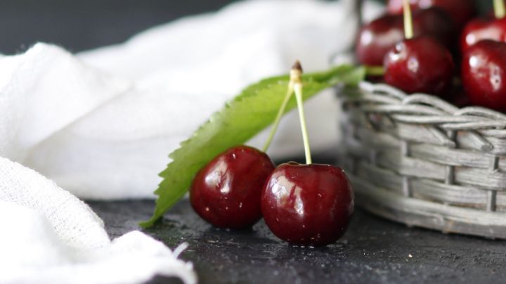 Saftige Kirschen liegen neben einem Korb auf einer wei&szlig;en Stoffserviette, die auf einer dunklen Oberfl&auml;che ruht.