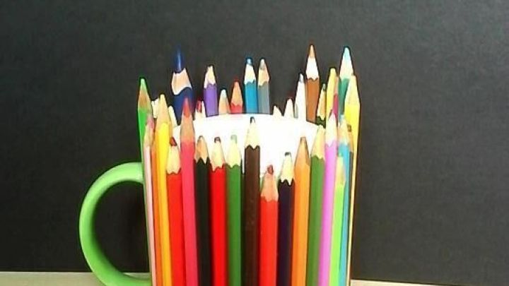 Bunte Stifte umh&uuml;llen Tasse, die auf einem Holztisch steht; der Hintergrund ist eine schlichte schwarze Wand.