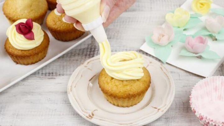 Eine Hand spritzt cremige Vanilleglasur auf einen Muffin, umgeben von weiteren Muffins und bunten Dekorationen.