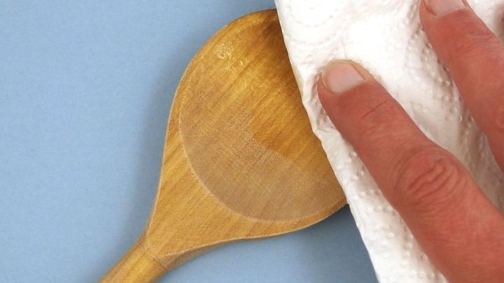 Ein Holzl&ouml;ffel wird mit einem Papiertuch abgewischt, um Speise&ouml;lreste auf einer blauen Unterlage zu entfernen.