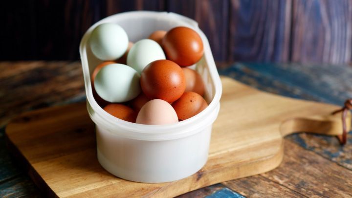 Ein Beh&auml;lter mit bunten Eiern steht auf einem Holzbrett, umgeben von rustikalen Holzfl&auml;chen.