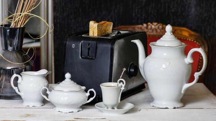 Ein Toaster steht auf einem Tisch und toastet Toastbrot, umgeben von einer eleganten Teekanne und Porzellangef&auml;&szlig;, mit getrockneten Pflanzen im Hintergrund.