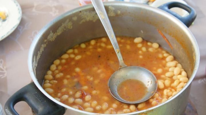 Wei&szlig;e Bohnen kochen in einem gro&szlig;en Topf, w&auml;hrend ein Kochl&ouml;ffel zum Servieren bereitliegt. Im Hintergrund sind Teller mit Beilagen sichtbar.