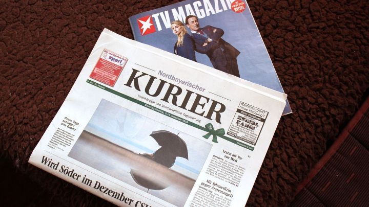 Eine Zeitungsauflage liegt auf einem Tisch, w&auml;hrend daneben ein TV-Magazin zu sehen ist, auf braunem Teppichboden.
