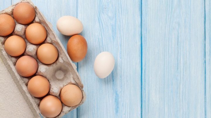 Ein Eierkarton mit braunen Eiern sowie zwei wei&szlig;e Eier liegen auf einer blauen Holzfl&auml;che, bereit zum Kochen.
