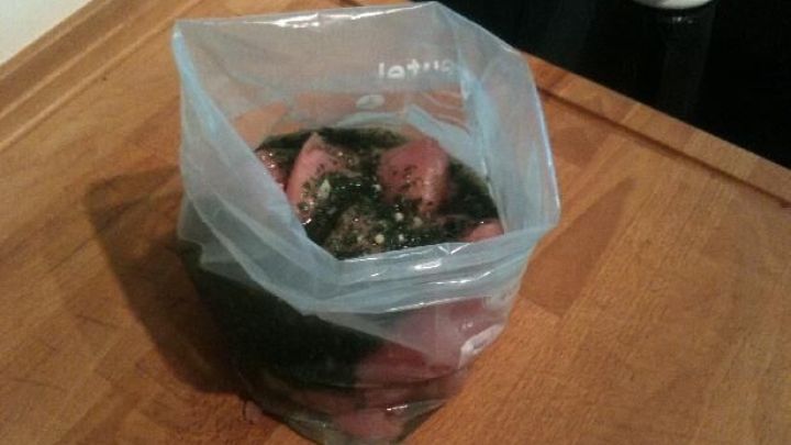 Ein Beutel mit Grillfleisch wird in Marinade auf einem Holztisch gezogen. Keine sichtbare Beschriftung.