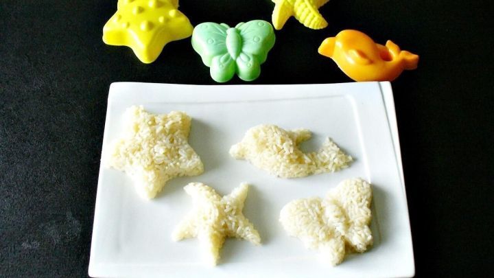 Reisfiguren auf wei&szlig;em Teller, geformt mit Sandkastenf&ouml;rmchen, die daneben liegen; schwarze Tischoberfl&auml;che.