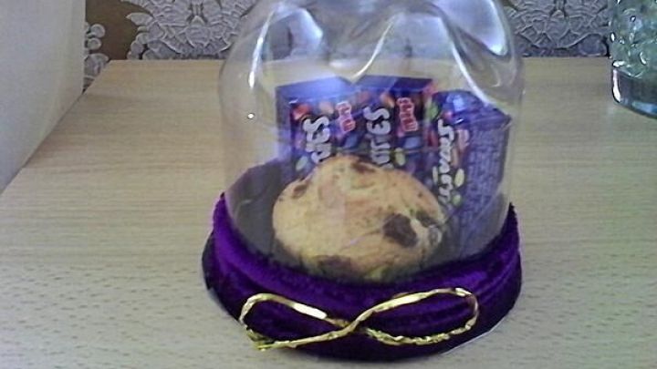 Pl&auml;tzchen unter PET-Flasche, umgeben von zwei Snack-Packungen, dekoriert mit violettem Stoff und goldener Schleife auf Tisch.