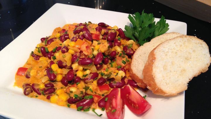 Buntes vegetarisches Chili mit Bohnen und Gem&uuml;se liegt auf einem wei&szlig;en Teller, w&auml;hrend frisches Brot daneben platziert ist.