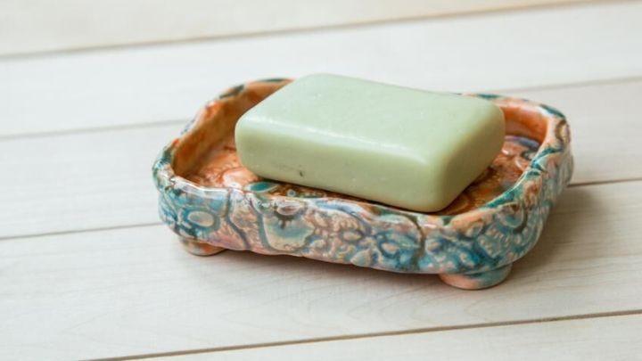 Eine gr&uuml;ne St&uuml;ckseife liegt ruhig auf einer dekorativen, handgemachten Keramikseifenschale auf hellem Holztisch.