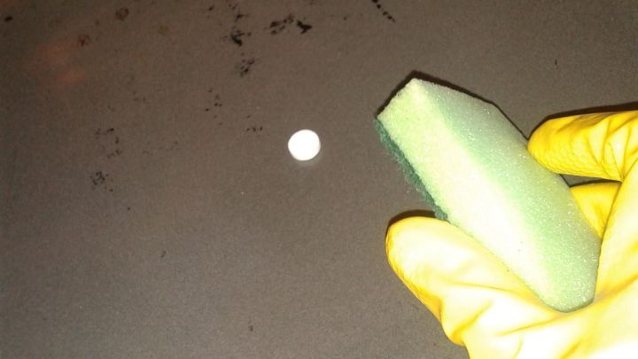 Eine gelb behandschuhte Hand h&auml;lt einen Schwamm, um Verkrustungen im Backofen auf einer schmutzigen Oberfl&auml;che zu entfernen.