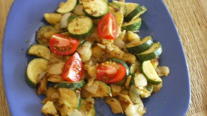 Eine bunte Kartoffel-Zucchinipfanne liegt auf einem blauen Teller und zeigt knusprige Kartoffeln, Zucchini und frische Tomaten.