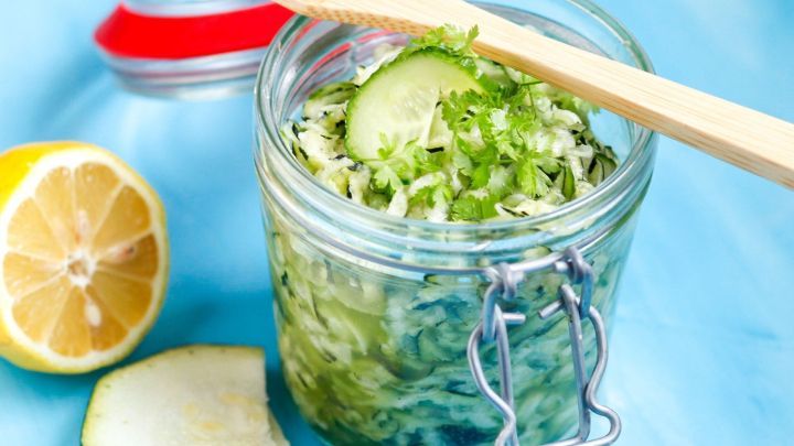 Ein Glas mit Gurken-Zucchini-Salat steht auf einem blauen Hintergrund, umgeben von Zitronenst&uuml;cken und einer Holzgabel.