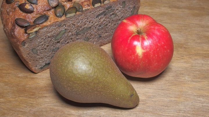 Ein dunkelbraunes Brot mit K&uuml;rbiskernen liegt neben einem roten Apfel und einer gr&uuml;nen Birne, die gesundes Abnehmen unterst&uuml;tzen.