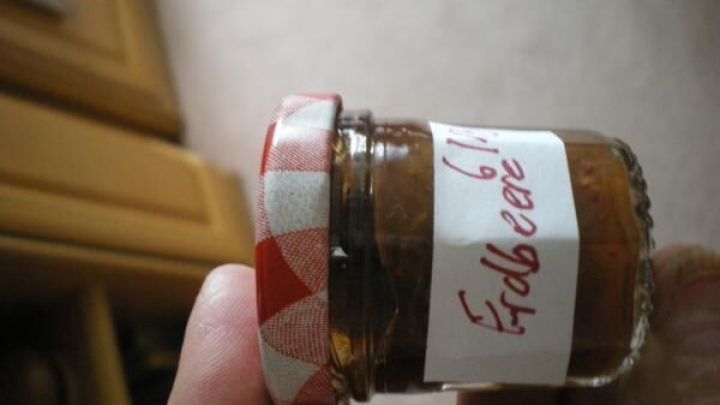 Miniglas mit Erdbeermarmelade wird in der Hand gehalten; Etikett beschriftet mit "Erdbeere 6.12.12". Regent thematisch.