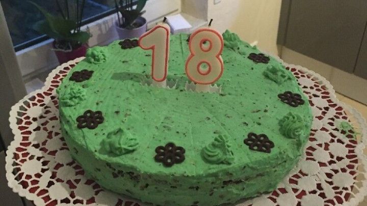 Torte mit gr&uuml;nem Minz-&Uuml;berzug, dekoriert mit Schoko-Blumen und "18"-Kerzen, wird auf einem Tortendeckchen gehalten.