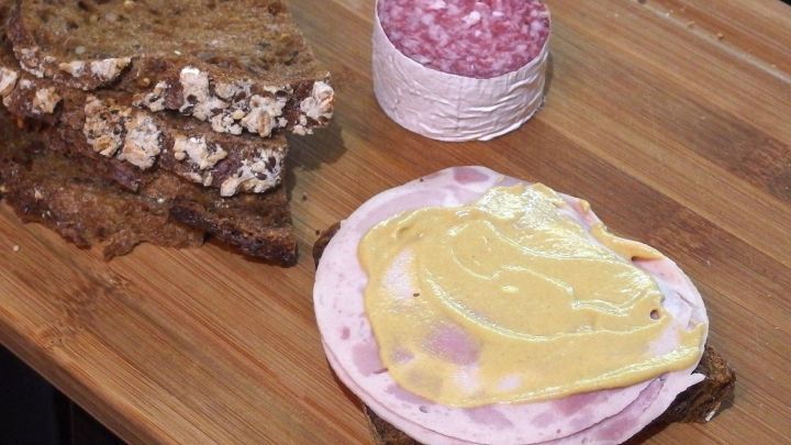 Ein belegtes Brot mit Schinken und Senf liegt auf einem Holzbrett, umgeben von dunklem Brot und Wurst.