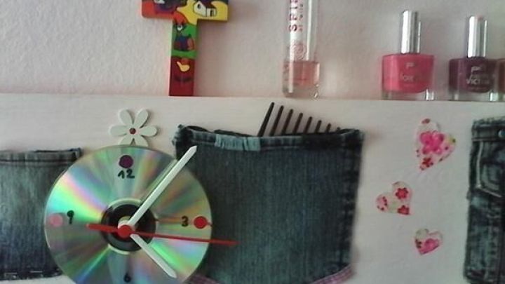 Eine CD-Uhr mit Zeigern ist auf einer Holztafel montiert, dekoriert mit einem bunten Kreuz, Nagellack und Jeansstoffen.