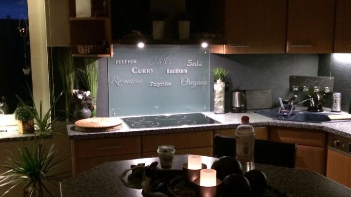 Ein K&uuml;chentisch mit Dekoration steht im Vordergrund, dahinter ein beleuchteter Fliesenspiegel mit den Worten "Pfeffer", "Chilli", "Curry", "Salz", "Basilikum", "Rosmarin", "Paprika", "Oregano" &uuml;ber einem Herd.