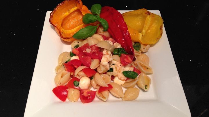 Nudelsalat mit Tomaten und Mozzarella liegt auf einem Teller, umgeben von gegrillten Paprikas und Basilikumbl&auml;ttern.