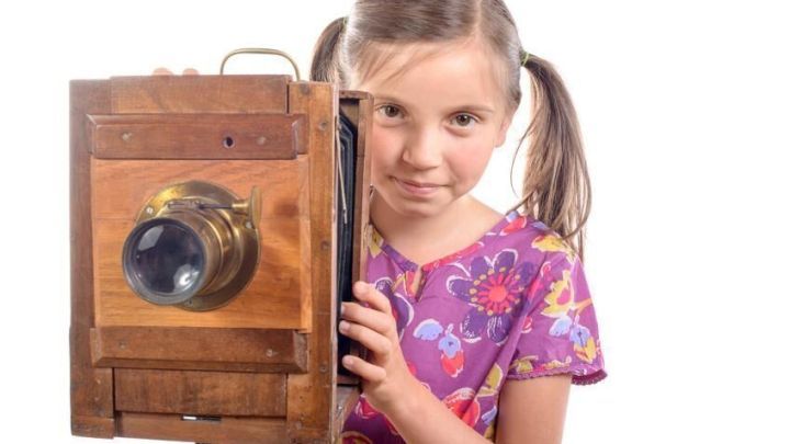 Ein M&auml;dchen h&auml;lt eine alte Holzfotokamera und schaut mit einem L&auml;cheln in die Kamera, bereit f&uuml;r ein Bild.