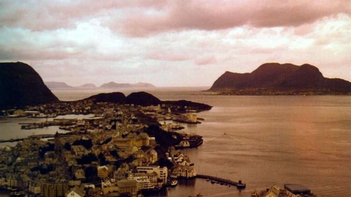 Ein K&uuml;stenpanorama zeigt eine Stadt mit Wasser und Bergen im Hintergrund, das Bild hat einen Rotstich.
