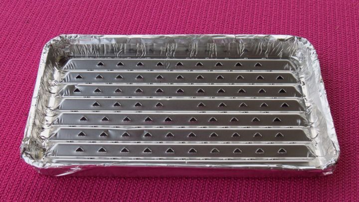 Eine Aluminium-Grillplatte liegt auf einem pinken Tisch, bereit zum Grillen im Elektroherd, ohne den Rost zu beschmutzen.