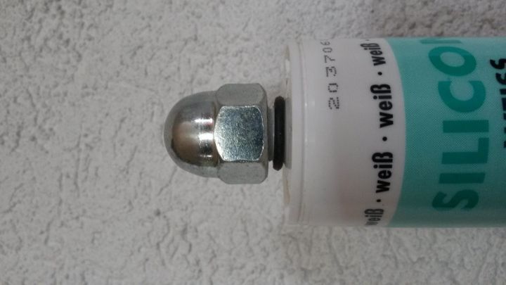 Eine Kartusche mit einem metallischen Verschluss steht auf einer hellen Wand. Der Verschluss sichert die Kartusche praktisch.