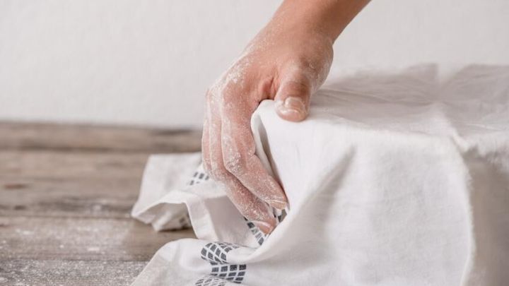 Eine Hand hebt ein Tuch an, das &uuml;ber einen Hefeteig gelegt ist, auf einer Holzoberfl&auml;che, bereit zum Gehen lassen.