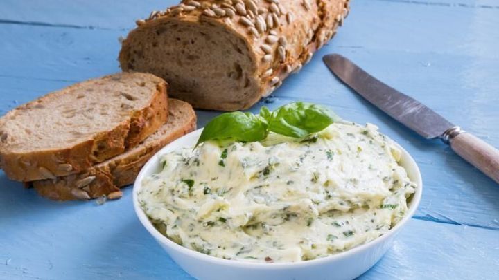 Eine Schale mit Kr&auml;uterbutter steht auf einem blauen Tisch neben einem Sonnenblumenkernbrot und einem Messer.