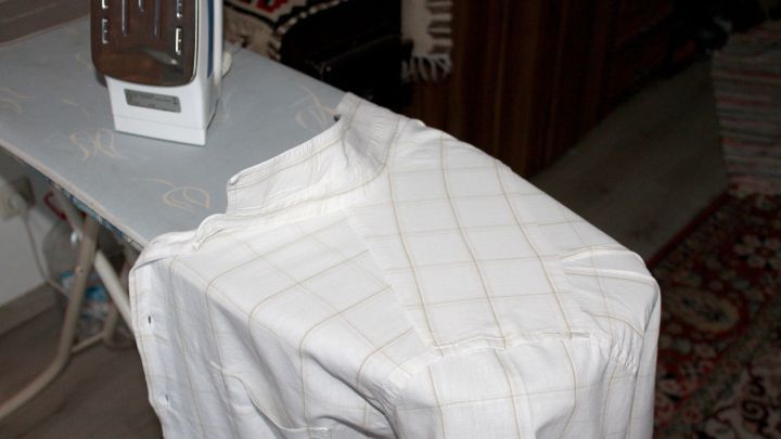 Ein Hemd liegt auf einem B&uuml;gelbrett, w&auml;hrend ein B&uuml;geleisen bereit zum B&uuml;geln daneben steht, in einem wohnlichen Raum.