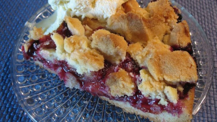 Eine k&ouml;stliche Zwetschgen-Streusel-Torte auf einem Glasteller, serviert mit einer Portion Sahne, auf einem blauen Tisch.