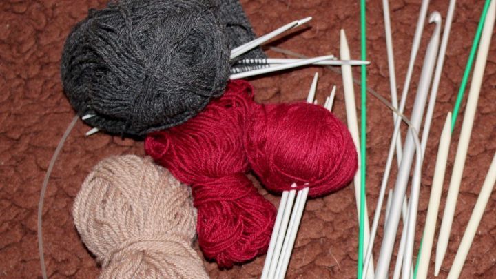 Graue, rote und beige Wollkn&auml;uel liegen neben gl&auml;nzenden Stricknadeln auf einem braunen Untergrund.