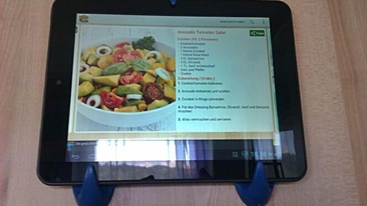 Ein Tablet ist mit Haken an der Wand montiert und zeigt ein Rezept mit einem bunten Salat. Die Umgebung wirkt k&uuml;chentechnisch.