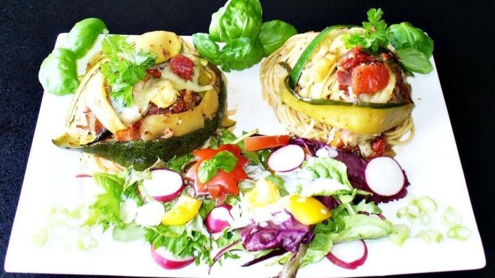 &Uuml;berbackene Zucchini-Nudelnester mit Schinken sind kunstvoll auf einem wei&szlig;en Teller angerichtet, umgeben von frischem Gem&uuml;se.