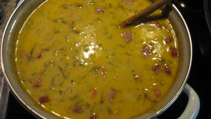 Eine gro&szlig;e Suppe mit einer cremigen, gelben Konsistenz kocht in einem Topf, verfeinert mit Petersilie und Schinken.
