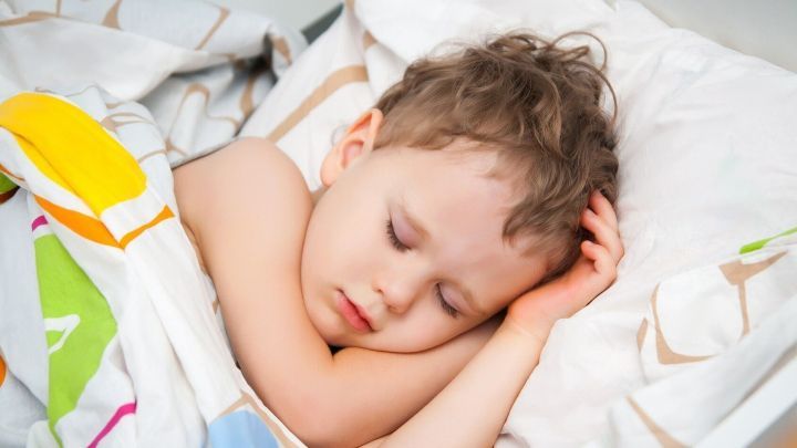 Ein Kind schl&auml;ft friedlich unter bunter Bettdecke in einem hellen Raum.