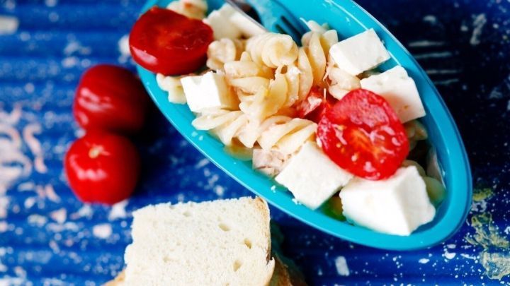 Ein bunter Nudelsalat mit Tomaten und K&auml;se liegt in einer blauen Schale auf einem Holzuntergrund, daneben liegt ein St&uuml;ck Brot.