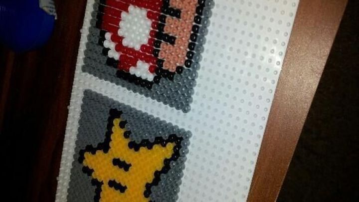 Bunte Untersetzer aus B&uuml;gelperlen zeigen einen Pilz und einen Stern auf einer wei&szlig;en Unterlage, die auf einem Tisch liegen.