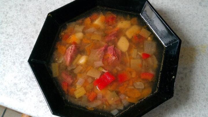 Sch&uuml;ssel mit klarer Suppe, gef&uuml;llt mit buntem Gem&uuml;se und Fleischst&uuml;cken, steht auf einem hellen K&uuml;chentisch.
