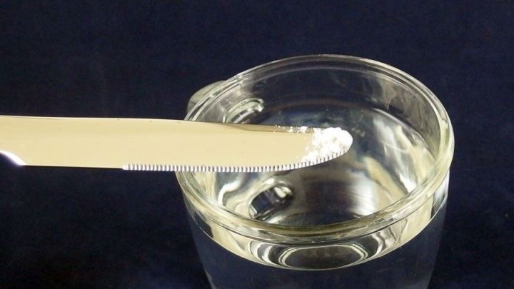 Ein L&ouml;ffel mit Backpulver steht &uuml;ber einem Glas Wasser, w&auml;hrend es darauf wartet, in die Fl&uuml;ssigkeit gegeben zu werden.