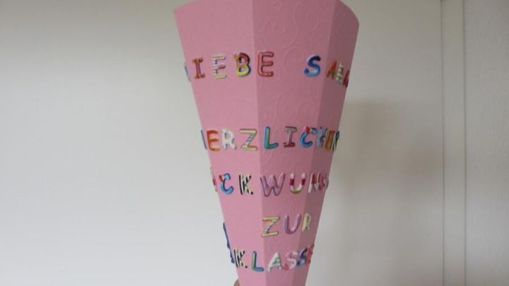 Eine pinke Mini-Schult&uuml;te wird mit bunten Buchstaben verziert, w&auml;hrend sie in einer Hand gehalten wird, vor einer Wand.