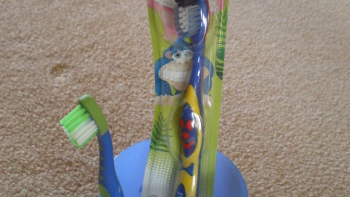 Eine blaue Kinderzahnb&uuml;rste steht in einem blauen Becher auf einem Teppich. Auf der Verpackung steht "KIDS 0-8".